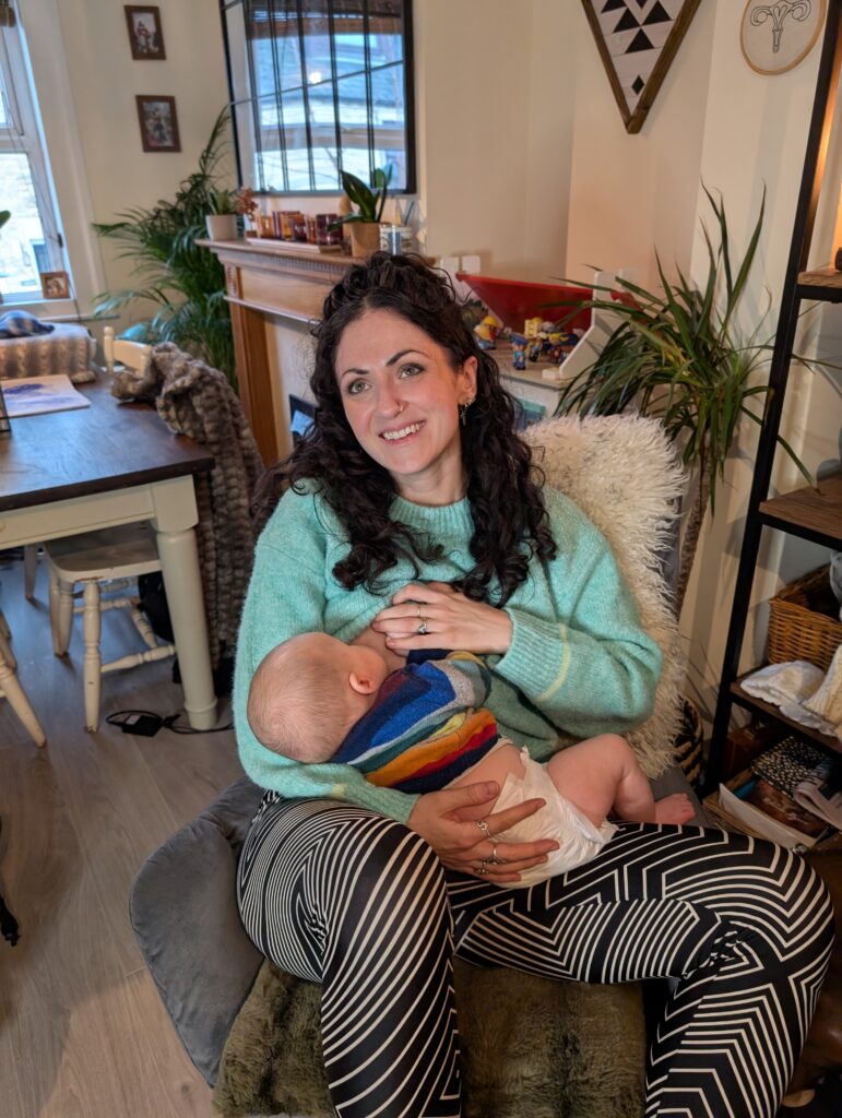 A woman sits comfortably in a cozy room, breastfeeding a baby while smiling, wearing a colorful outfit.